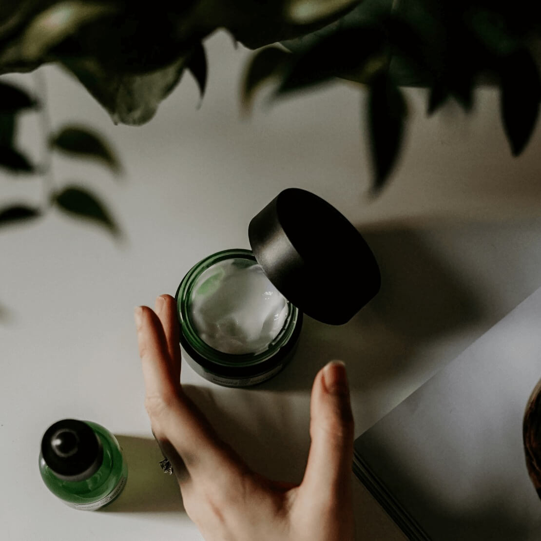 A hand holding a jar of luxury skincare among green leaves